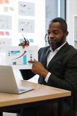 Fototapeta premium An African American man in a suit wearing a headset points to a bar graph showing upward growth during a virtual meeting