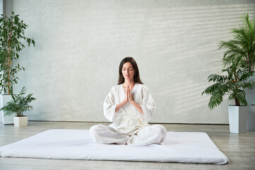 Beautiful woman in white dress sitting on mat in lotus position before Thai massage, practicing...