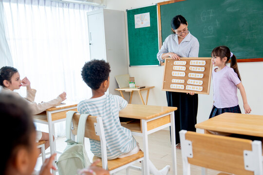 The teacher hold chart of multiplication table and tell little girl to present about this chart for her friends in front of the classroom.