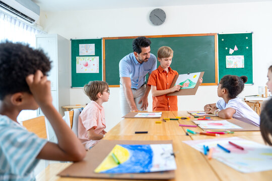 Wide shot of the teacher stand beside and try to make a boy with blond hair present his drawing or painting in front of the classroom and also ask the idea or the meaning of the picture.