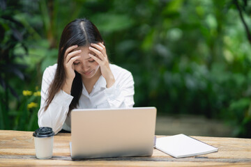 Young professional woman with stress headache sitting at a desk with laptop, coffee cup, and notebook in a serene green outdoor environment