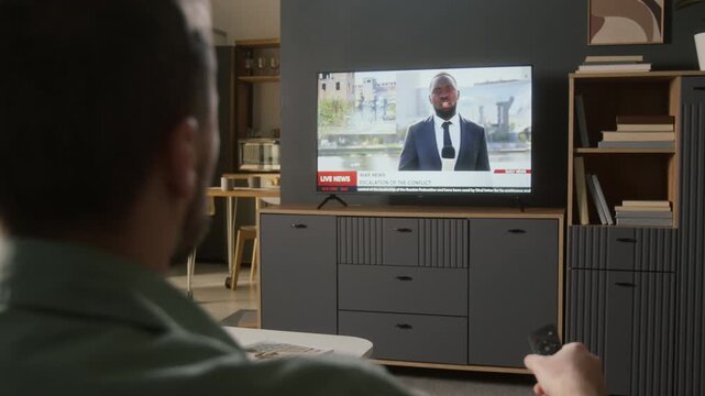 Over-shoulder rear shot of man watching TV news broadcast with African American journalist or politics expert commenting on military war conflict, footage of soldiers raiding ruins and firing guns