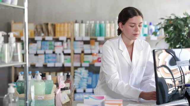 Qualified female pharmacist works at a computer in a European pharmacy. Pharmacist checks the availability of goods in a spreadsheet against the background of shelves with medicines. High quality 4k