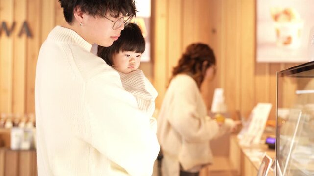 Soft light fills a cozy bakery. Woman holds a curious child close. Another woman browses pastries thoughtfully. Warm tones embrace the scene. Perfect for family moments or cozy cafe imagery