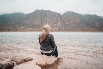 Woman in puffer jacket and cap sits by a calm lake with misty mountains in the background © Jezabel