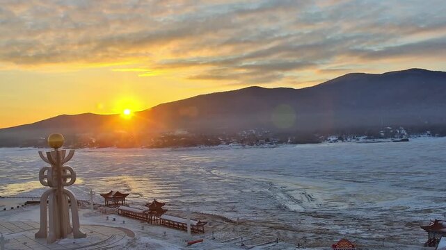 A beautiful winter sunrise over the frozen Amur River in Fuyuan, featuring traditional pavilions and a snowy mountain landscape.