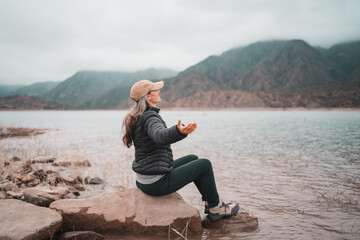 Woman in puffer jacket and cap meditating by a serene lake with misty mountains © Jezabel