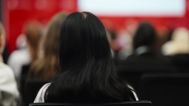 Back view of a dark-haired attendee seated among a blurred audience in a red-walled conference hall, listening intently to a live presentation during a professional seminar