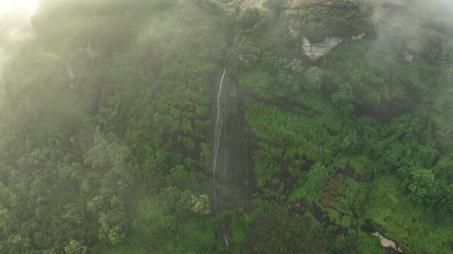 A cinematic lateral drone movement reveals a slender waterfall flowing down a lush green hillside, partially veiled by soft morning mist and low-hanging clouds in west java
