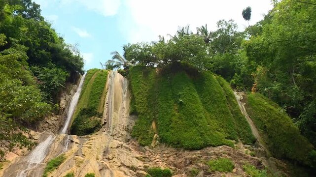 A cinematic horizontal panning movement reveals the unique Curug Koja waterfall, featuring water cascading over tiered limestone rock formations in a lush West Java forest