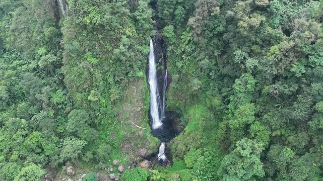 A wide aerial establishing shot of a majestic single-drop waterfall hidden in the dense tropical mountains of Cianjur, west Java