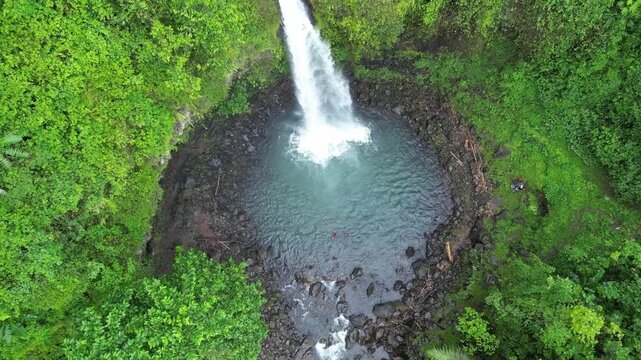 A high-angle overhead drone shot looking down at people swimming in the natural turquoise pool of Ketemu Jodoh waterfall