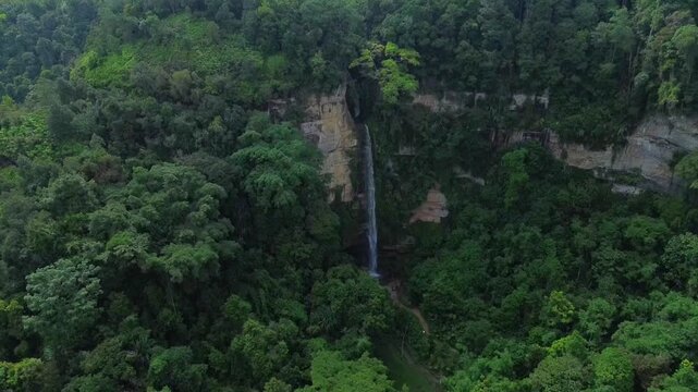 A cinematic aerial drone shot moving toward to Sarasah Tanggo waterfall as it drops from a limestone cliff into a lush, dense tropical rainforest in West Sumatra, Indonesia