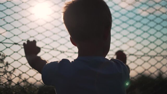 Boy touching chainlink fence with both hands outdoors at sunset. Child presses against wire barrier silhouette. Boy stands at fence touching mesh. Child hands on chainlink fence with sunlight.