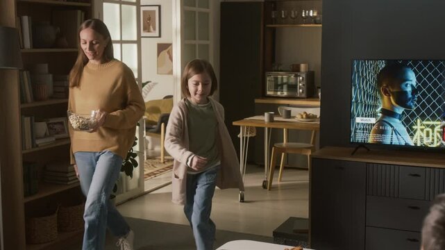 Full shot of excited young Caucasian girl, mother and pet dog walking out of kitchen with bowl of popcorn, sitting down on couch to join father, to watch movie on TV with online streaming service
