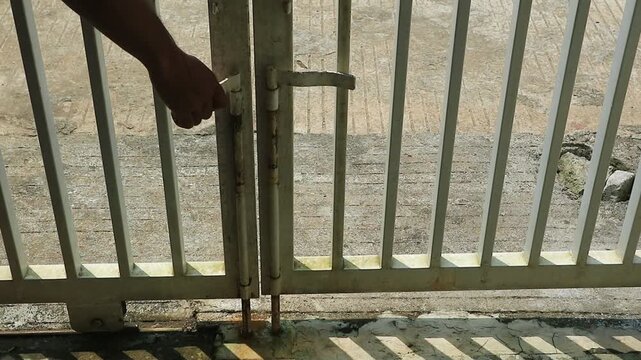 Close up footage of a person hand pulling down a locking lever on metal gate showing everyday security action with visible texture of iron bars and outdoor ground surface