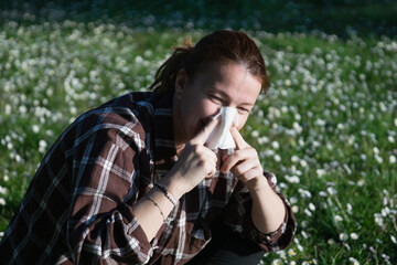 woman blowing nose in daisy field brown plaid shirt seasonal allergy suffering looking away lying on green grass in sunny park with copy space for banner design concept of healthcare, medicine, nature © Lana Pietukhova