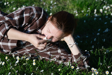 woman blowing nose in daisy field brown plaid shirt seasonal allergy suffering looking away lying on green grass in sunny park with copy space for banner design concept of healthcare, medicine, nature © Lana Pietukhova