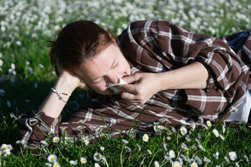 woman blowing nose in daisy field brown plaid shirt seasonal allergy suffering looking away lying on green grass in sunny park with copy space for banner design concept of healthcare, medicine, nature © Lana Pietukhova