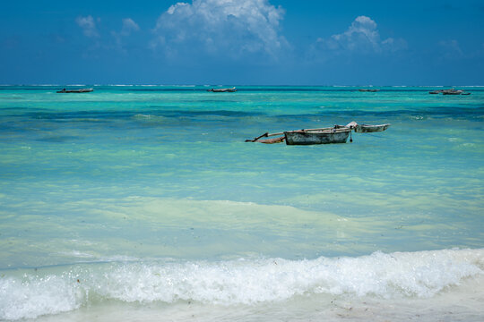A single traditional wooden boat floats on calm, multi-toned turquoise ocean water under a clear blue sky in Zanzibar, Tanzania.