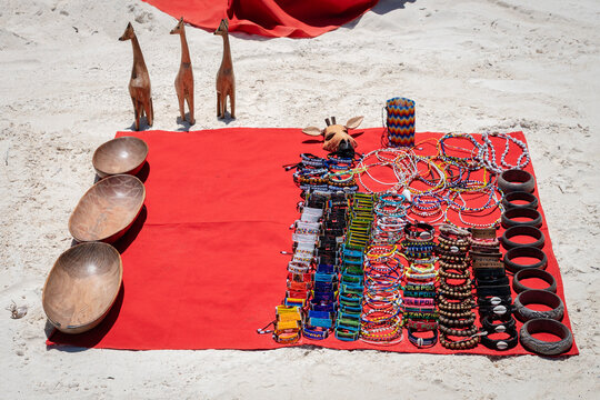 Traditional African handmade jewelry, beaded bracelets, wooden bowls, and giraffe figurines displayed on a red cloth on a white sand beach in Zanzibar, Tanzania.