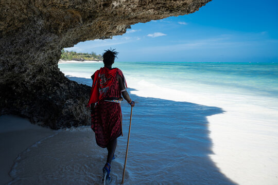A Maasai man in traditional red clothing walks along a pristine white sand beach under a dramatic overhanging coral rock formation in Zanzibar, Tanzania.
