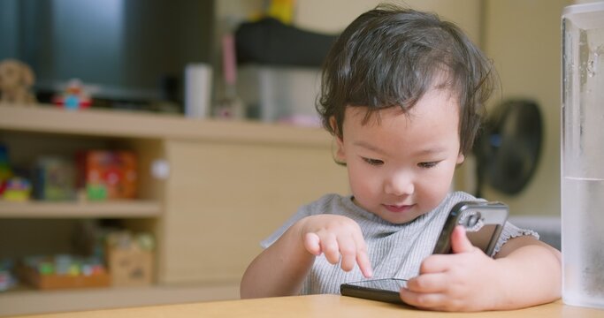 Focused Asian toddler taps and swipes a smartphone screen at a home living room table, focusing on early digital literacy, screen time engagement, and modern childhood technology habits.