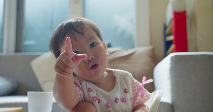 Playful Asian toddler points index finger toward the camera lens inside a bright living room, focusing on interactive communication, early childhood development, and joyful expression.