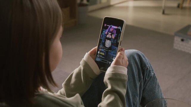 Over-shoulder shot of young girl watching female cosplayer with cat ears, purple wig and kawaii outfit performing fun dance on social media live stream, while relaxing on couch at home