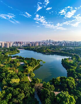An aerial view showcases a serene lake winding through a lush green park, bordered by a sprawling cityscape under a vibrant blue sky dotted with fluffy white clouds.
