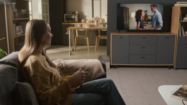 Medium full side shot of young Caucasian woman sitting down on couch with bowl of chips, watching love drama on flat screen digital TV, couple violently arguing and fighting