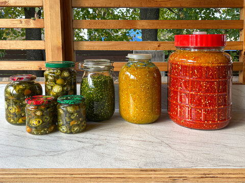 Colourful jars of pickled vegetables on wooden table, home canner arranging harvest display with cucumbers, olives, peppers in brine, glass lids catching