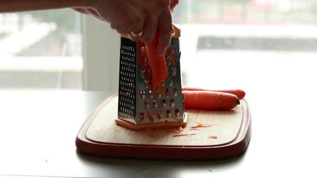 Close-up of woman's hands grating a fresh carrot on a stainless steel box grater in natural light