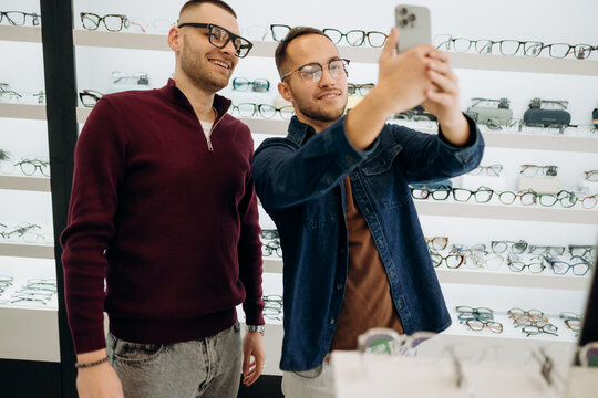 Two friends smile as they take a selfie in an optical store. They are trying on glasses in front of shelves filled with eyewear. It is afternoon and the store is well-lit.