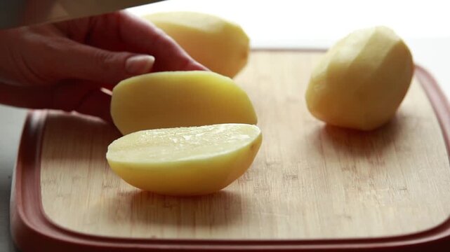Close-up of woman's hands slicing and peeling fresh raw potatoes on a rustic wooden cutting board in bright natural light