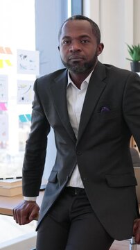 A confident African American man in a suit smiles warmly while leaning on a desk in an office setting with charts on the wall