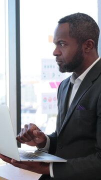 An African American businessman in a suit looks intently at his laptop screen while standing near a window with a city view