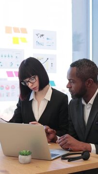 A woman in glasses points at a laptop screen while discussing data with an African American man in a suit during a business meeting