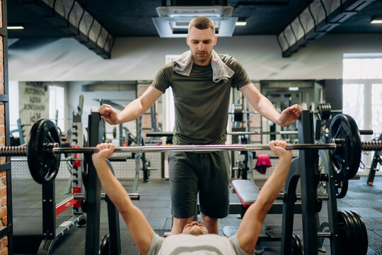 Two men are in a gym. One man is lifting weights on a bench press. The other man spots him to ensure safety while he lifts. They are focused on the exercise.