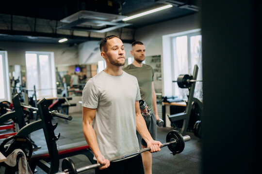 Two men perform strength training with weights inside a gym. One man holds a barbell while the other stands nearby with a dumbbell. Sunlight enters through windows.