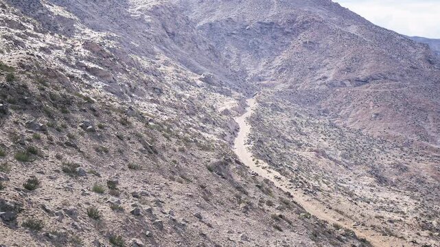 A winding dirt road cuts through rough, rocky mountainous terrain under a bright sky. Sparse vegetation grows among the rocky outcrops, suggesting a dry climate.