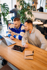 Two men are focused on a laptop screen at a cafe during the day. They are sitting at a wooden table with a tablet and notebooks nearby while discussing something on the computer.