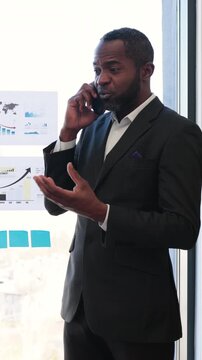 A concerned executive gestures while discussing charts and financial reports during a mobile call in a bright, glass-walled office