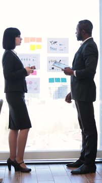 Two professionals, an African American man and an Asian woman, collaborate on a project using charts and sticky notes on a glass wall