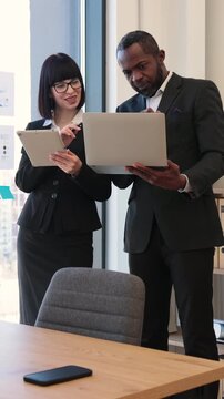 Two professionals consult a laptop and tablet in a modern office by a window while preparing a presentation for a meeting with colleagues and stakeholders