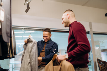 Naklejka premium Two men are choosing clothes in a store. One man holds a jacket while the other smiles and shows interest. They are looking through different styles and colors. The scene captures their enjoyment.