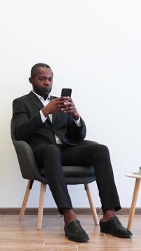 An African American man dressed in a formal suit sits in a modern chair while intently focused on his mobile phone