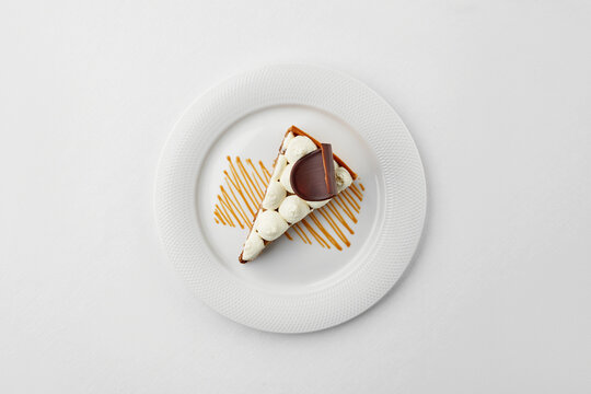 Top view of a gourmet tart slice with whipped cream, chocolate garnish, and caramel drizzle on a white plate. Minimalist aesthetic on a white background.