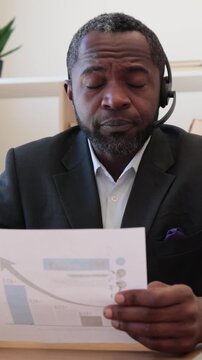 An African American man wearing a headset and suit examines a document with a rising graph, indicating business growth and analysis