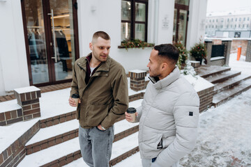 Two men are outside a building in winter. They are walking and talking while holding coffee cups. Snow covers the ground and the surroundings look cold.